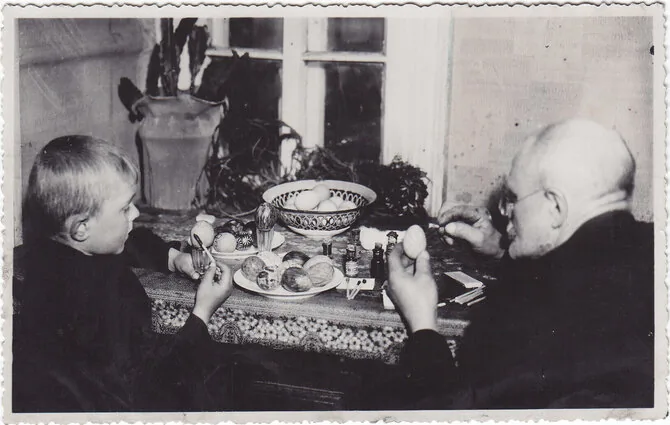 Pranešimo autorių nuotr. /Fotonuotrauka. Senelis (dešinėje) su anūku (kairėje) dažo velykinius margučius prie uždaroje patalpoje esančio stalo. XX a. 3–4 deš. (Trakų muziejus, CC BY)